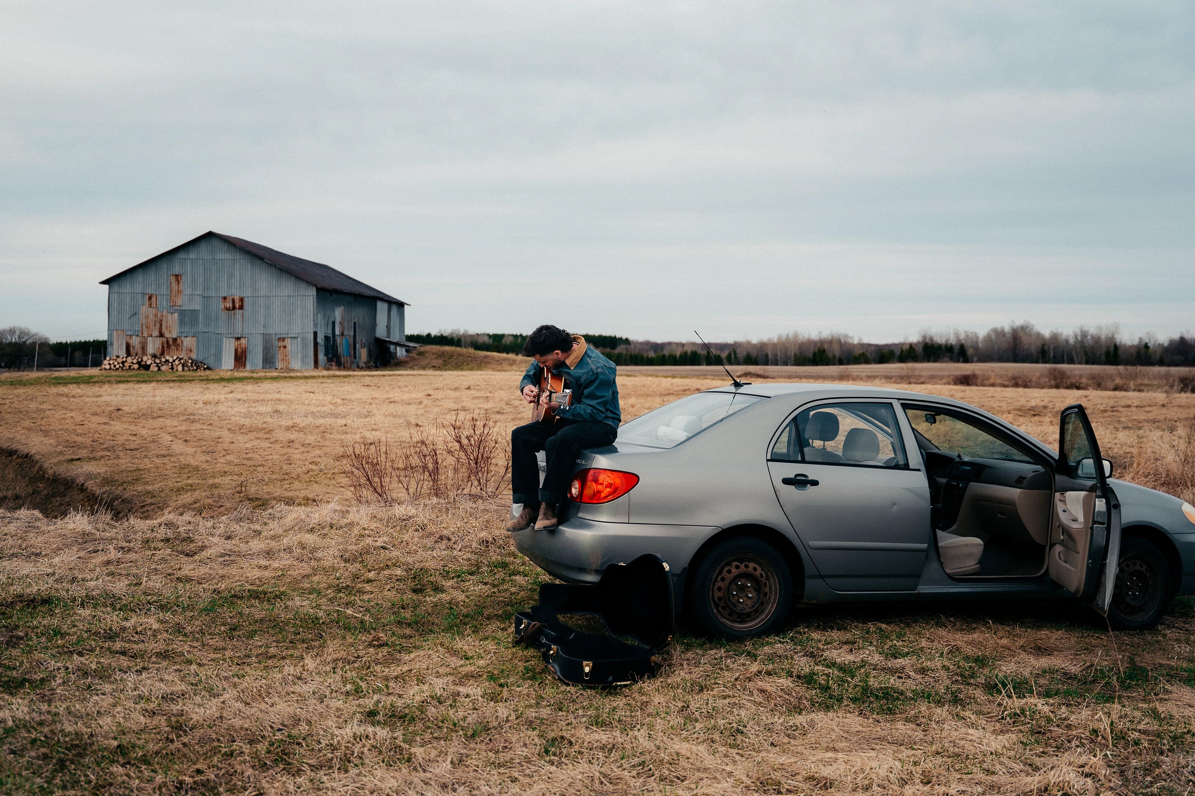 Joseph Poirier joue de la guitare, assis sur le coffre d'une voiture au bord d'un champ, une grange au loin